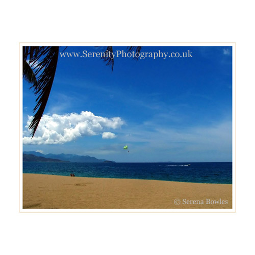 Blue skies, sand, sea. A couple sit on the beach watching a paraglider.